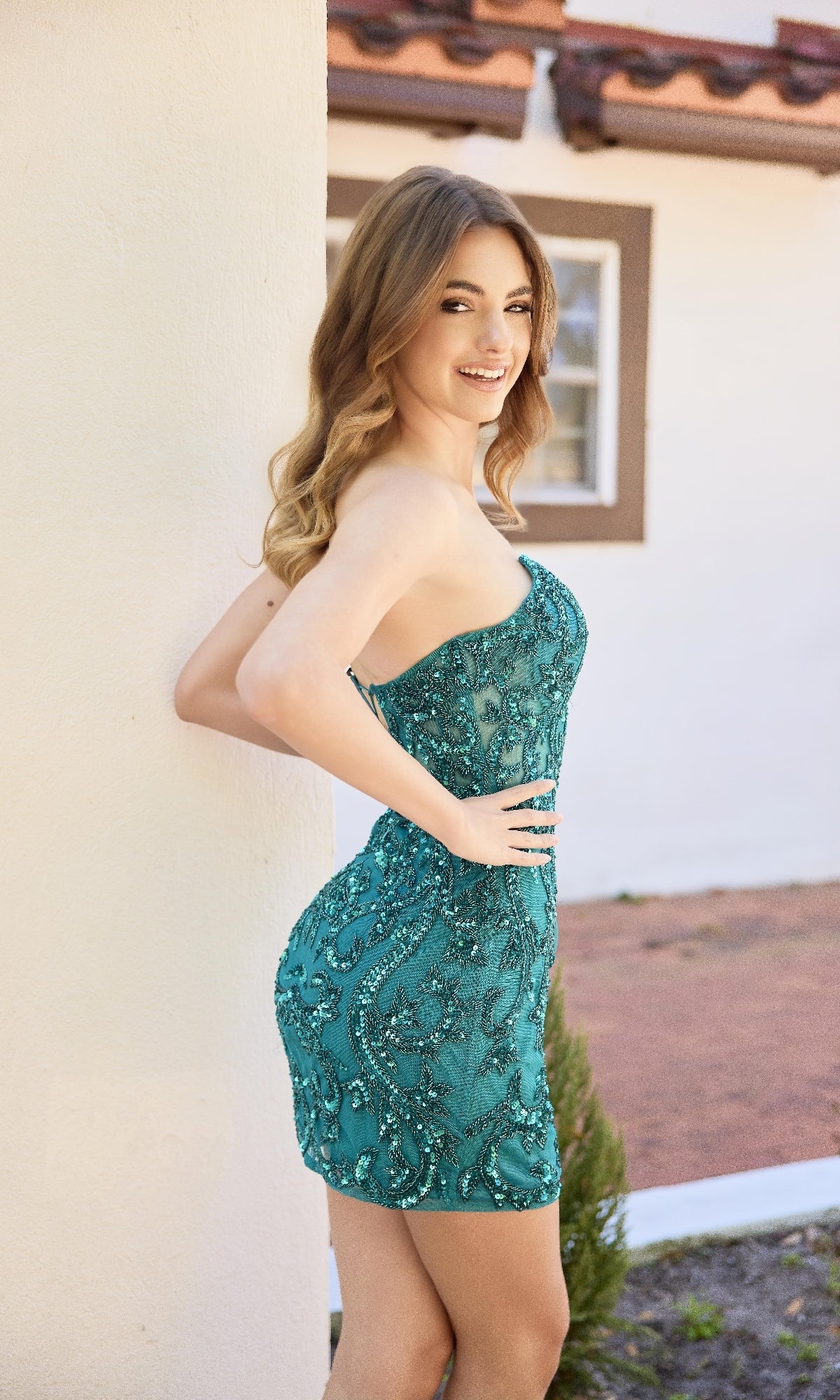 A woman poses outdoors in the Strapless Short Sequin Hoco Dress: Primavera 4430, leaning against a white wall. The short sequin cocktail dress features a lace-up back, with a building and pathway in the background.